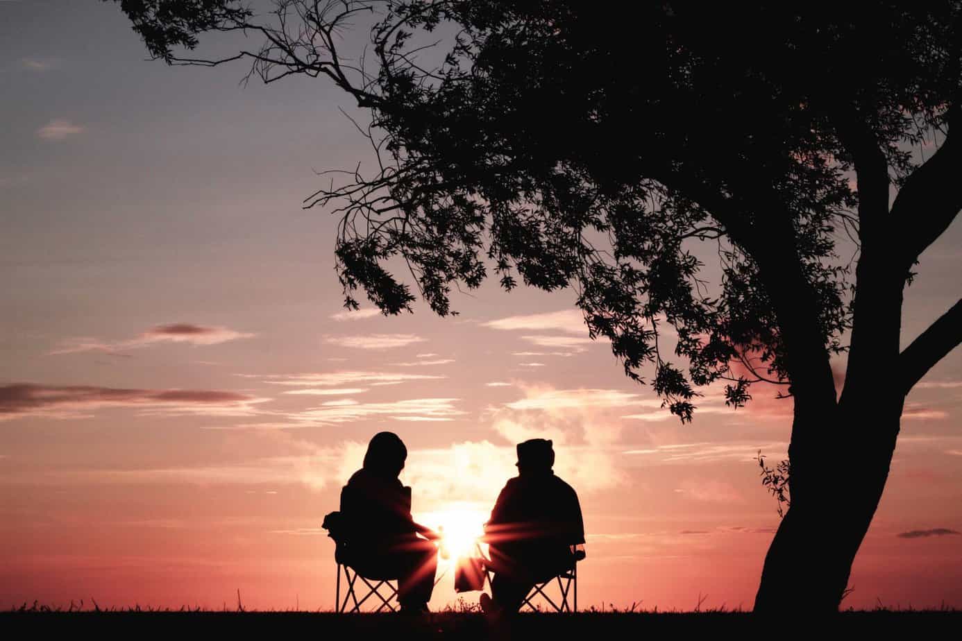 shadow of Two people sitting on lawn chairs outside under a tree talking and watching the sunset