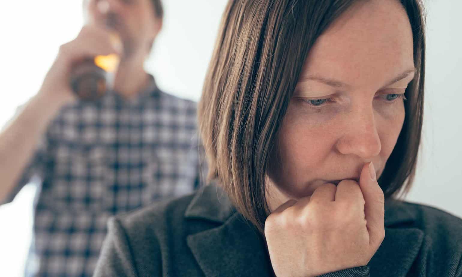 woman looks upset as husband takes a drink of beer in background