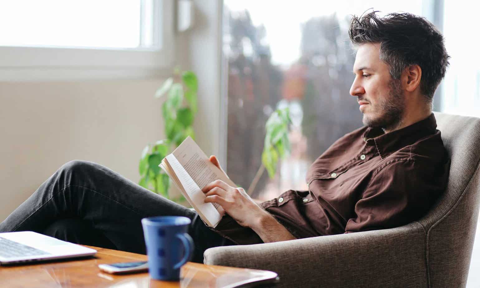 Man reading while sitting on chair by desk with mug laptop and phone on wooden table