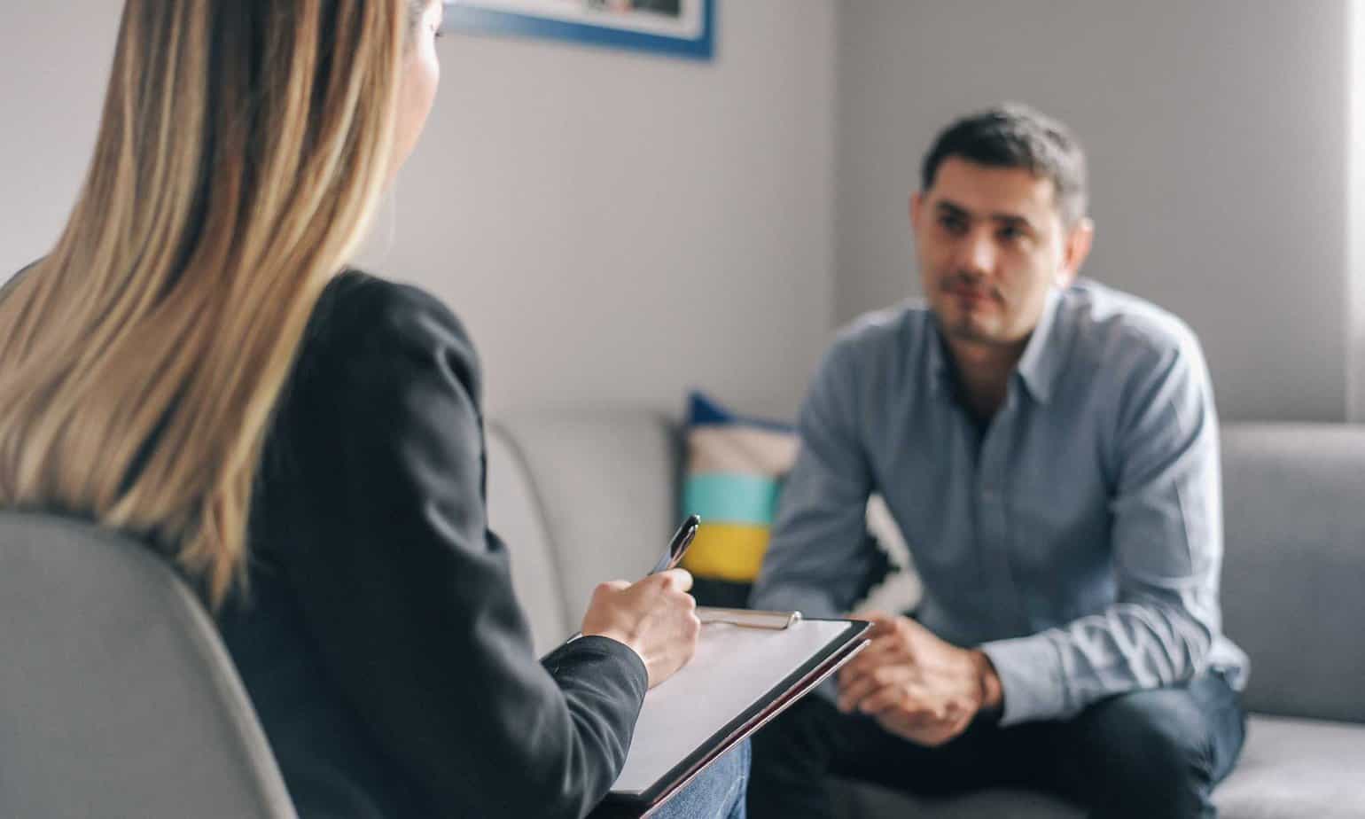 Man sitting on couch across from blonde female therapist taking notes on clipboard