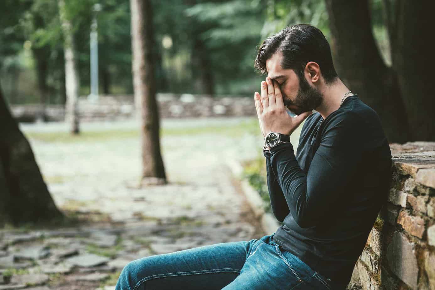 Man in long black shirt and jeans sitting on park bench outside near trees with hands on head in prayer pose after a relapse