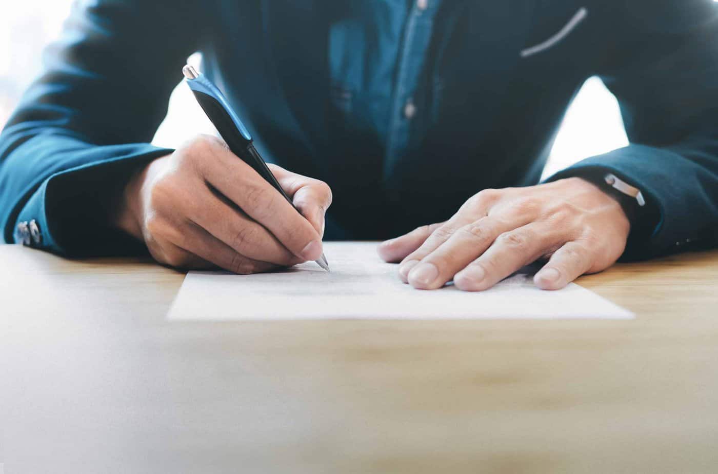 close up of man in blue suit with hands writing a letter on a piece of paper with a pen