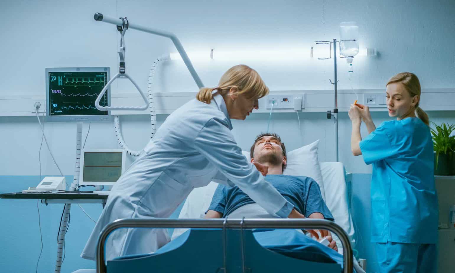 Female Doctor and Nurse check vitals on unconscious male patient in blue hospital room with medical equipment