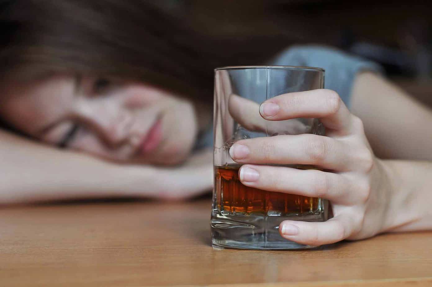 woman with depression lies on table clutching a glass containing alcohol