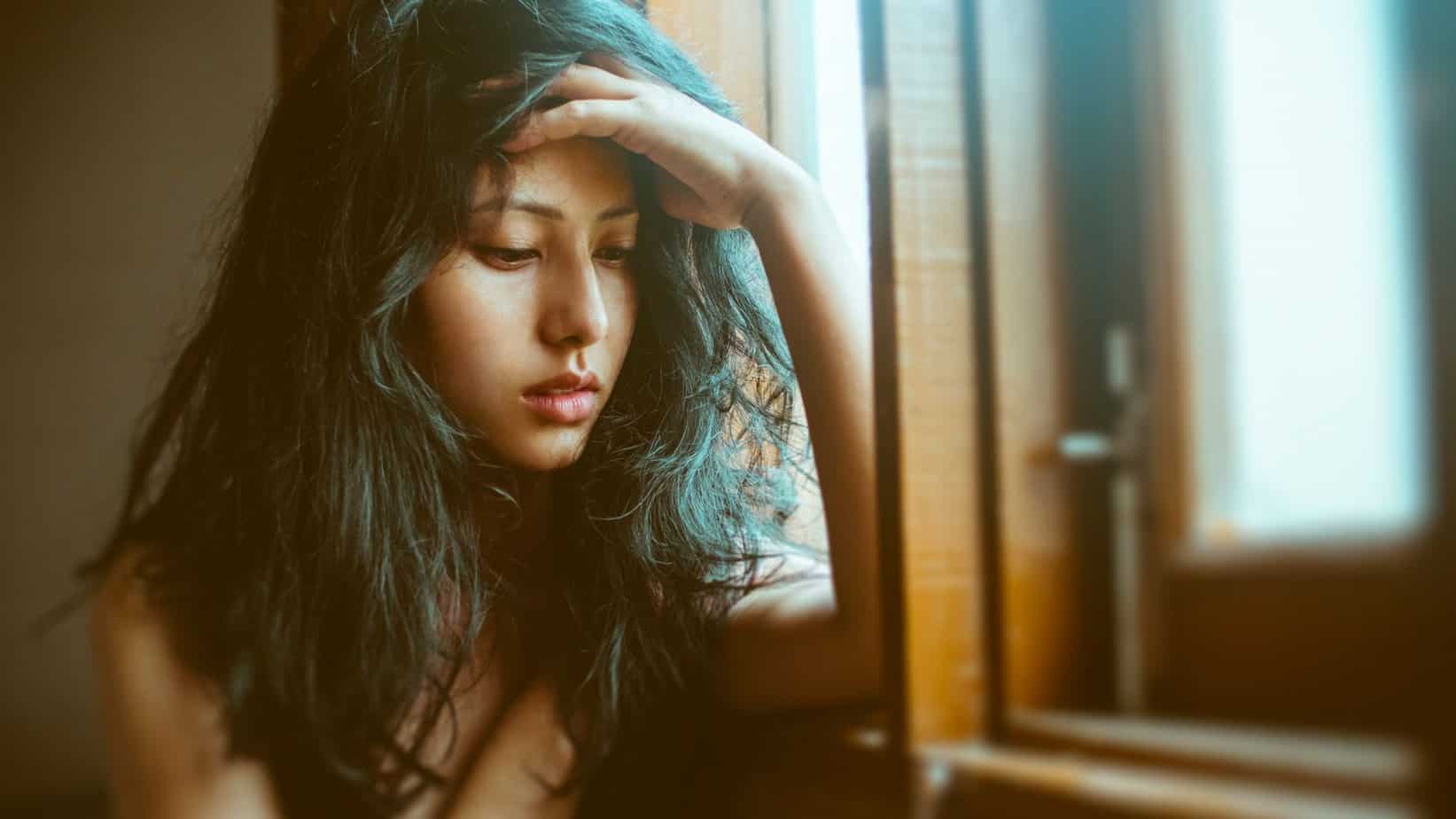 Dark haired woman with hand on head looking down and sad by sill of window