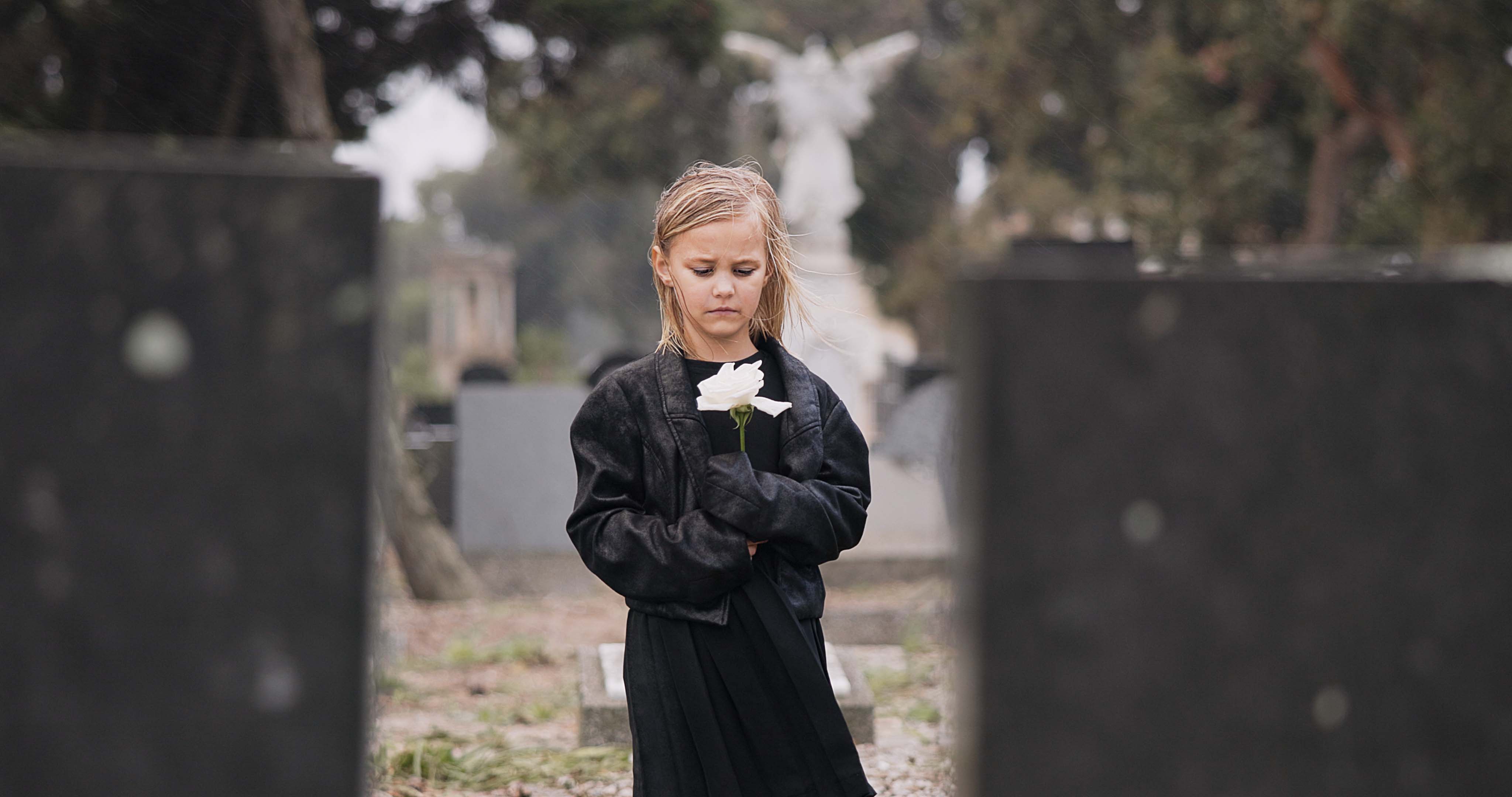 Young girl standing at a grave while holding a white flower.