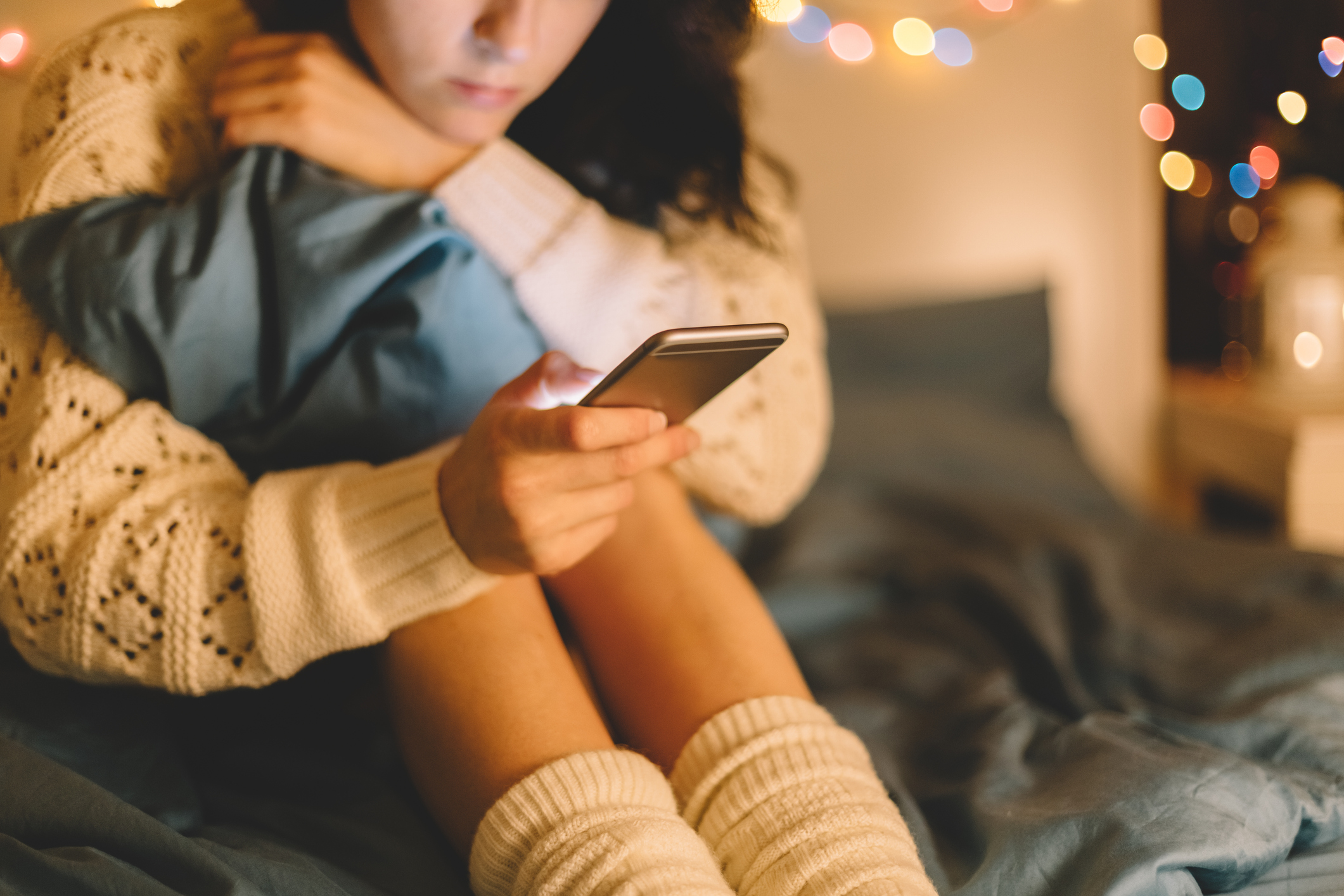 A girl is sitting on her bed with her legs curled up, scrolling on her phone.